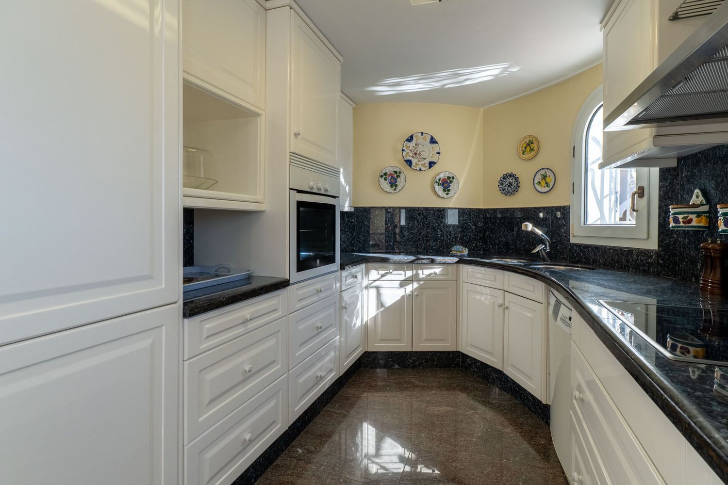 Independent kitchen featuring white paneled cabinets, dark granite countertops and backsplash. It includes a built-in oven, sink, and an arched window providing natural light.