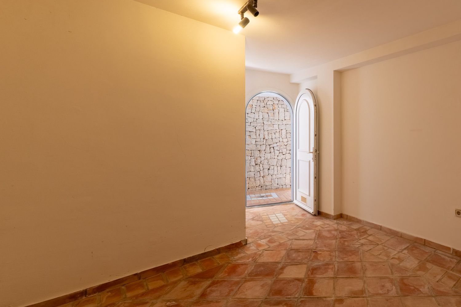 Interior hallway featuring terracotta tiled flooring and an arched white door opening to an outdoor patio with an irregular stone wall. Ceiling spotlights.