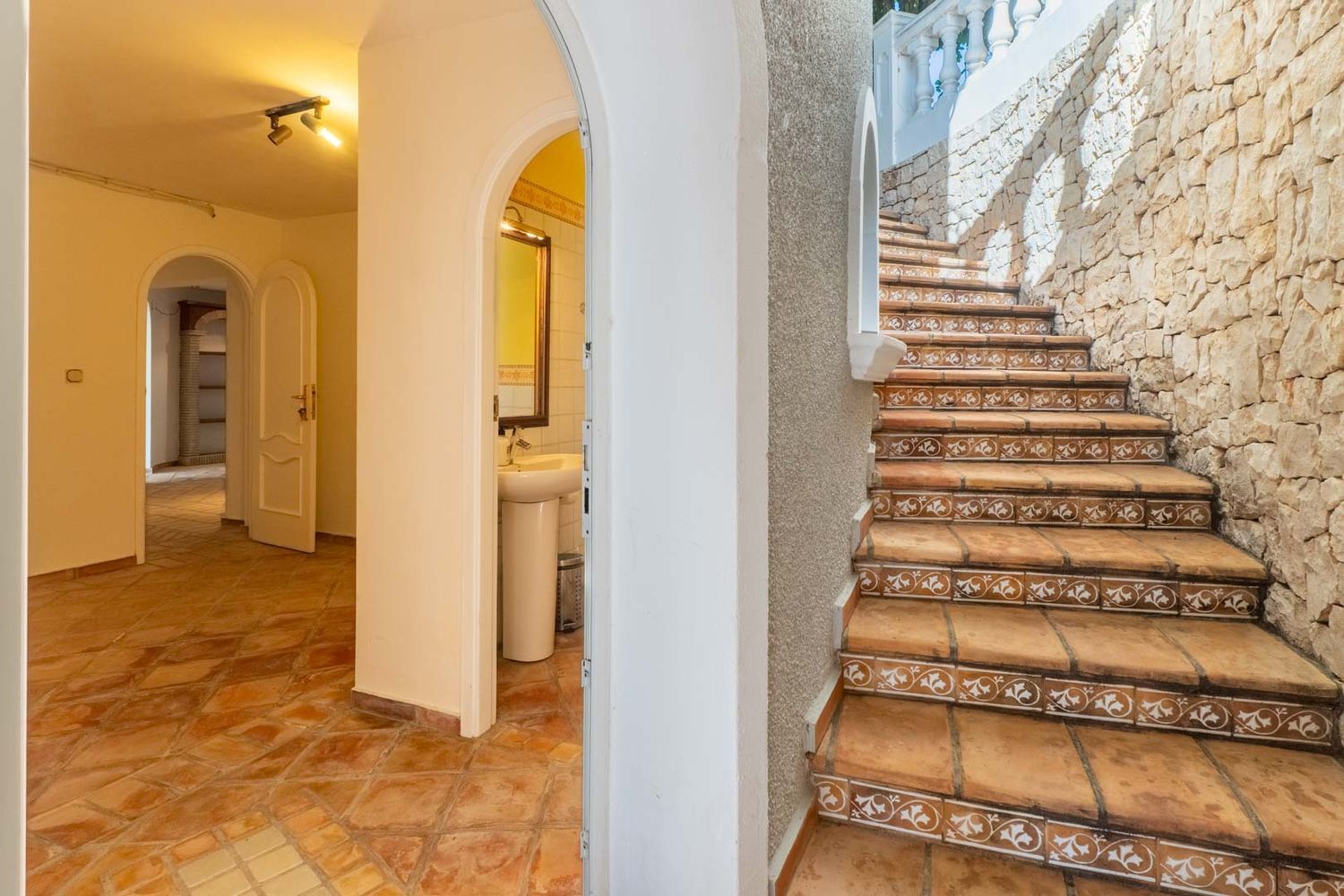 Interior hallway with terracotta tiled floors and arched entrances, contrasting with an exterior stone staircase featuring decorative risers and a robust stone wall.