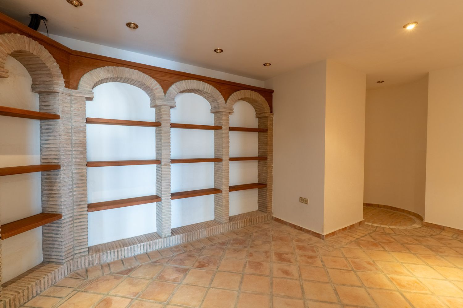 Interior room featuring terracotta floor tiles and a built-in shelving unit with brick arches and wooden shelves. Recessed lighting illuminates a distinct corner.