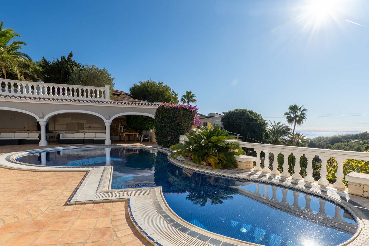 Irregularly shaped blue mosaic pool with terracotta tiling, adjacent to a covered arched terrace with white outdoor sofas. Distant sea views visible through lush greenery.