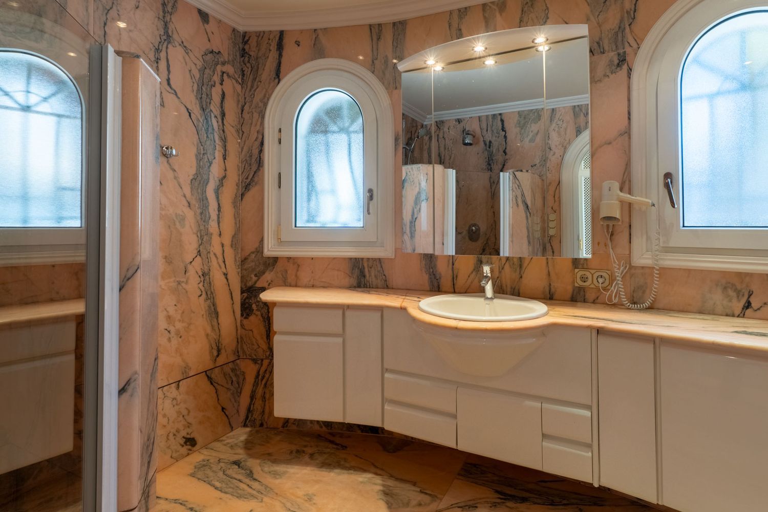 Main bathroom clad in veined pink marble, featuring a white floating vanity with integrated sink and two arched frosted windows.