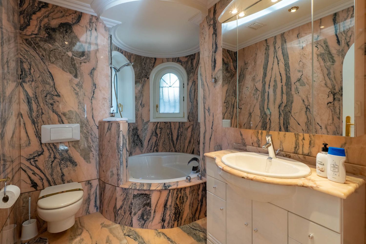Main bathroom featuring pink and black veined marble, a built-in bathtub in a curved alcove, and an arched window providing natural light.