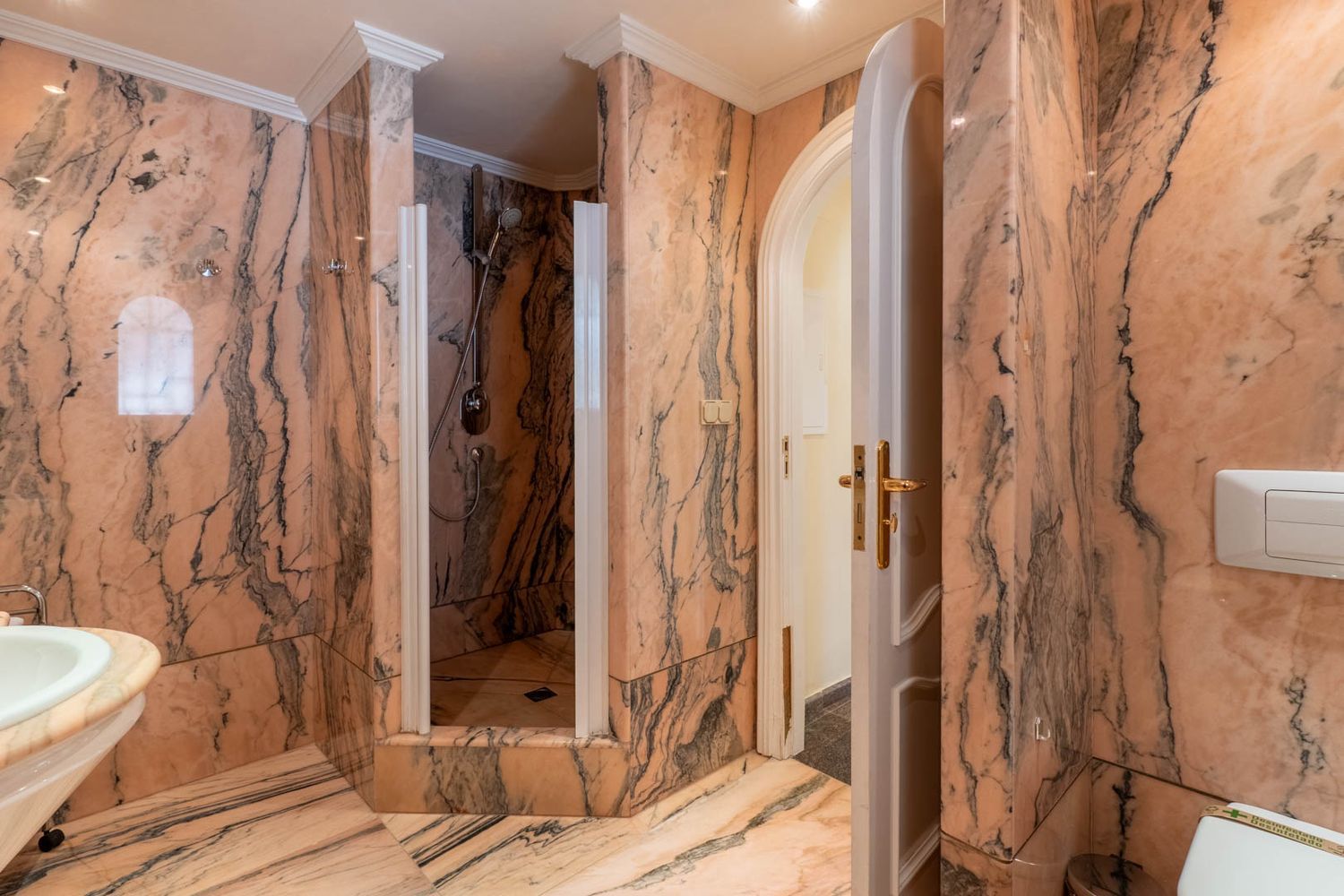 Main bathroom fully clad in veined pink marble, featuring a walk-in shower with a glass screen and an arched doorway.
