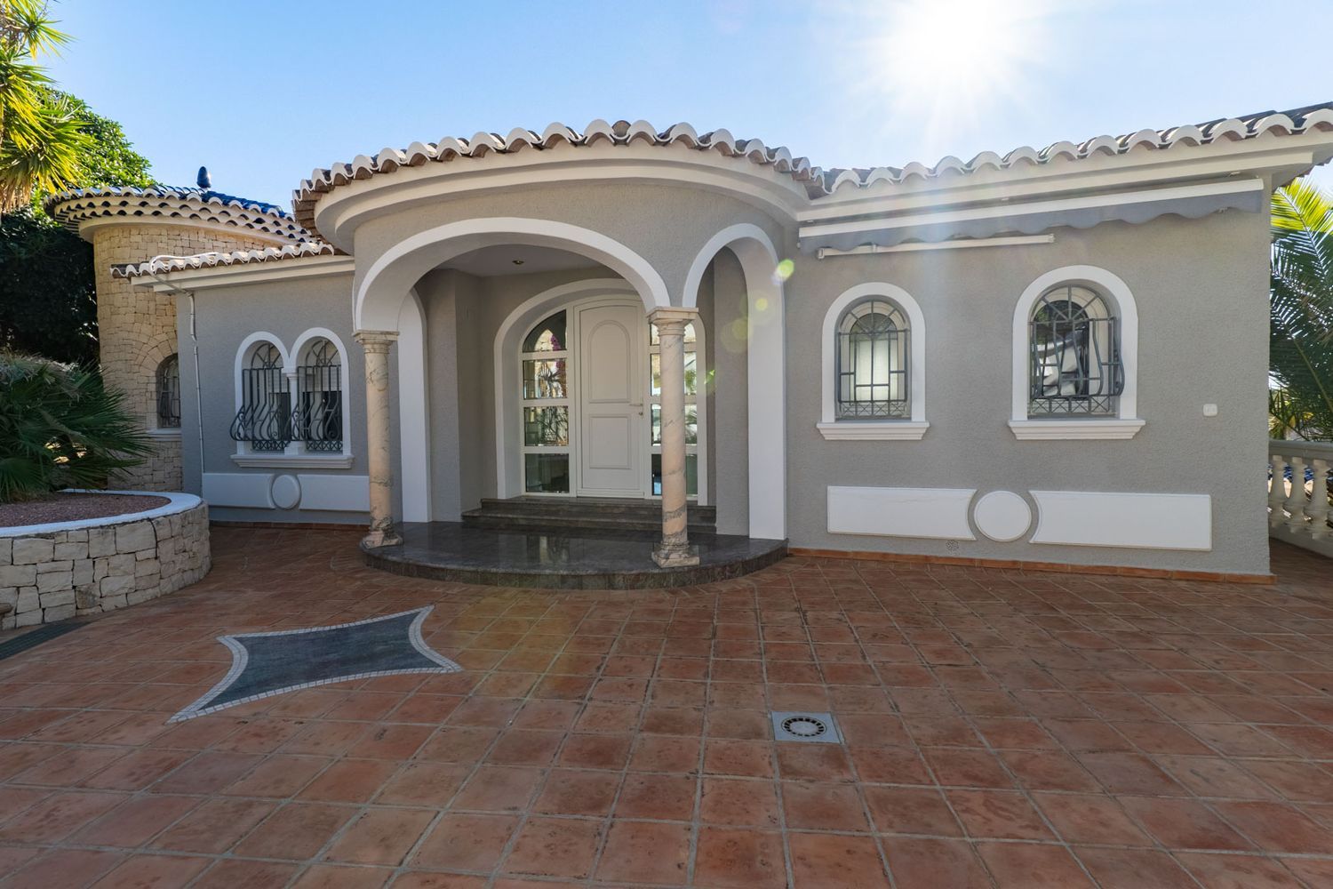 Main entrance of the villa featuring a curved porch with marble columns and white arches. Arched windows with decorative wrought iron grilles and terracotta paving.