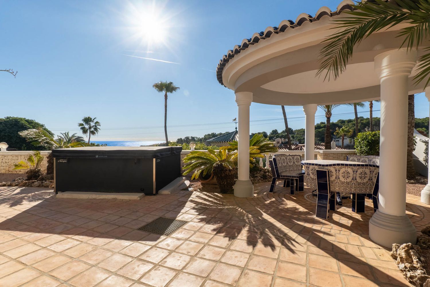 Outdoor terrace featuring a hot tub and a covered dining area with white columns and a tiled roof. A blue and white tiled table and chairs sit on terracotta flooring, offering sea views.