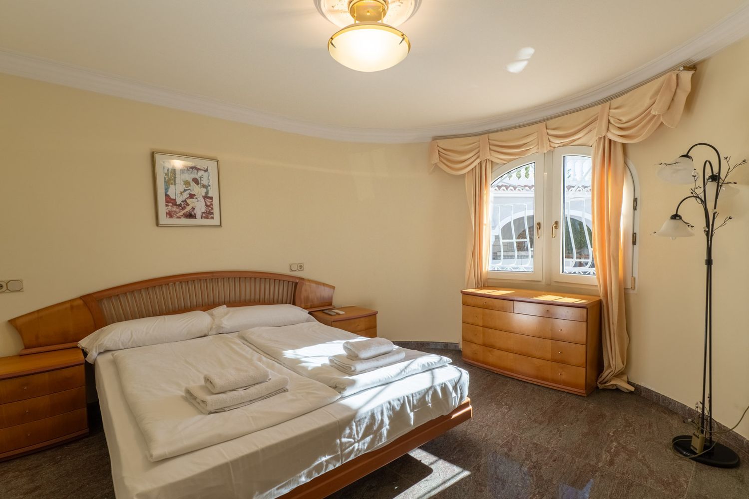 Spacious bedroom featuring a double bed with a wooden headboard, matching nightstands, and a dresser. Natural light enters through a curved window, illuminating the dark tiled floor.