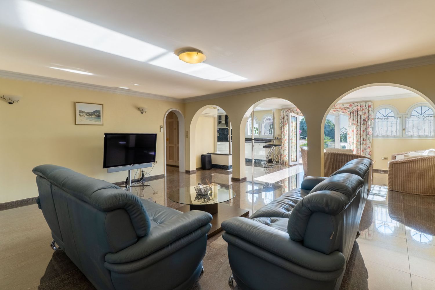 Spacious living area featuring glossy dark marble floors, leather sofas, and arched openings leading to the kitchen and outdoor terrace. Natural light fills the room.