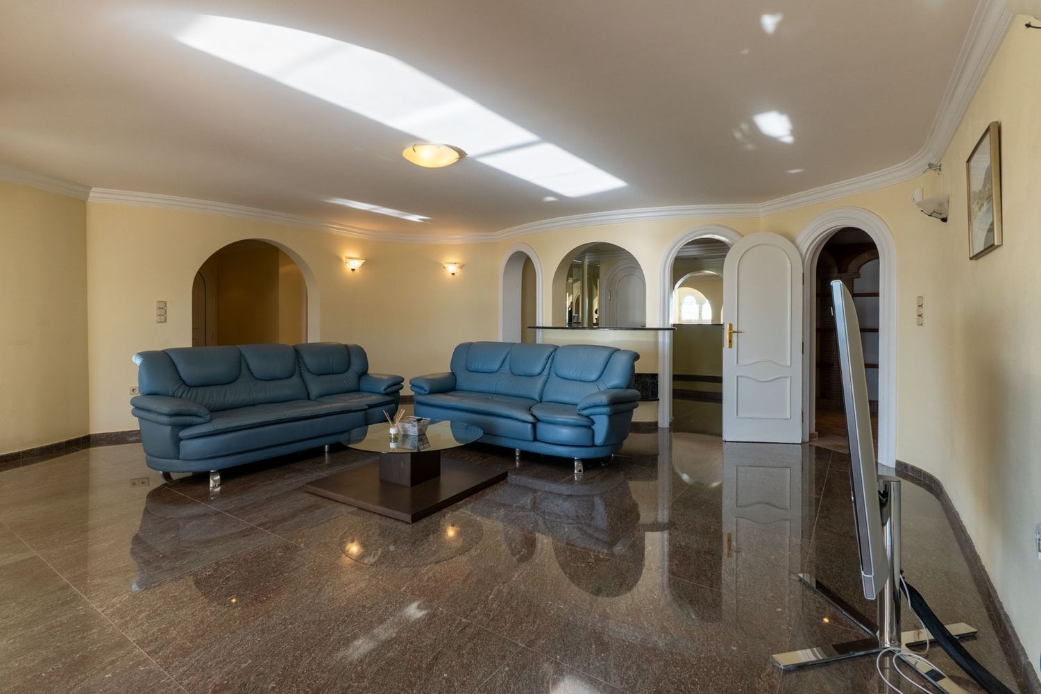 Spacious living area featuring polished dark marble floors, blue leather sofas, and multiple arched openings leading to adjacent rooms. Natural light reflects on the ceiling.