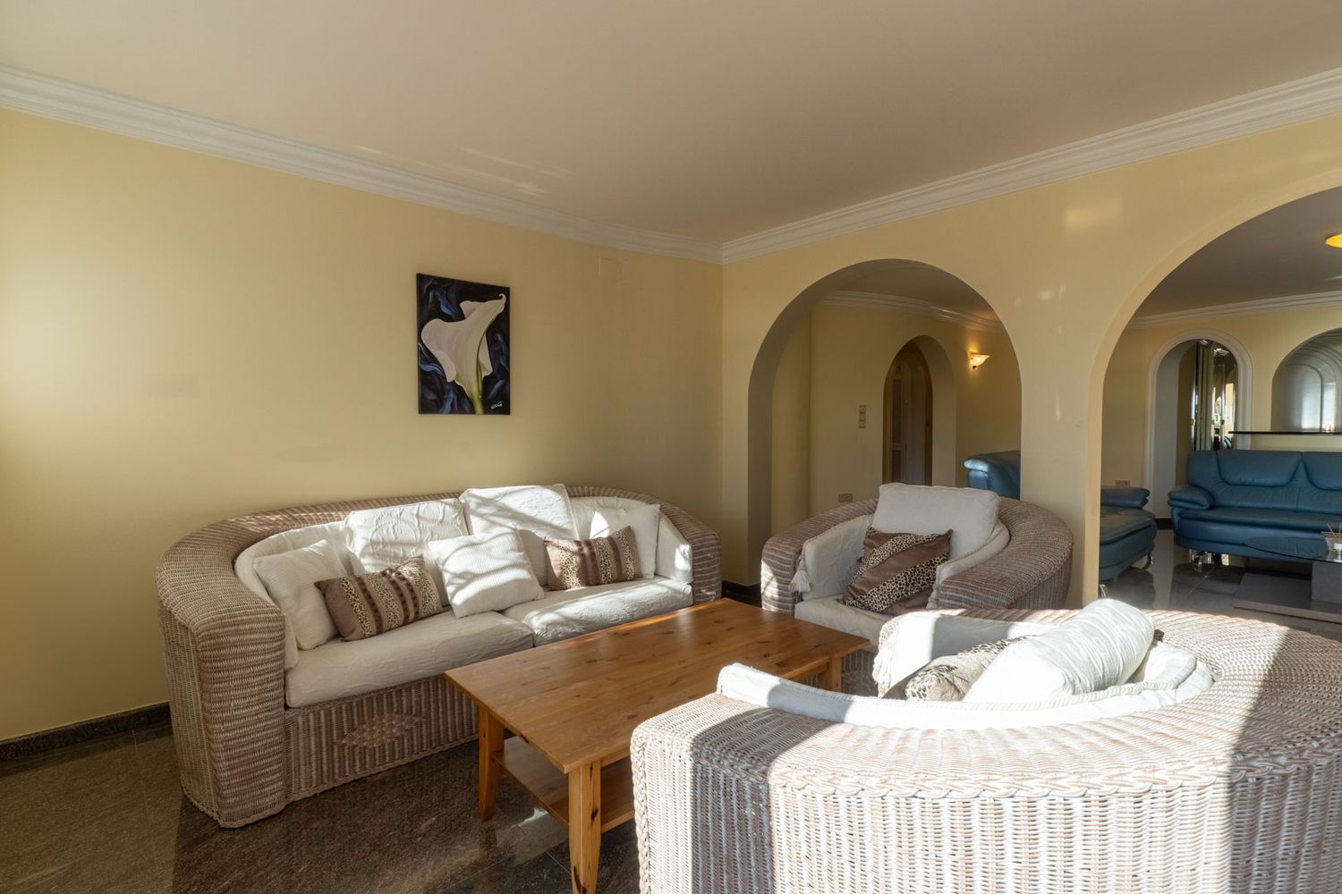 Spacious living area with wicker sofas, light cushions, and a wooden coffee table on dark marble flooring. Elegant arches connect to an adjacent room, bathed in natural light.