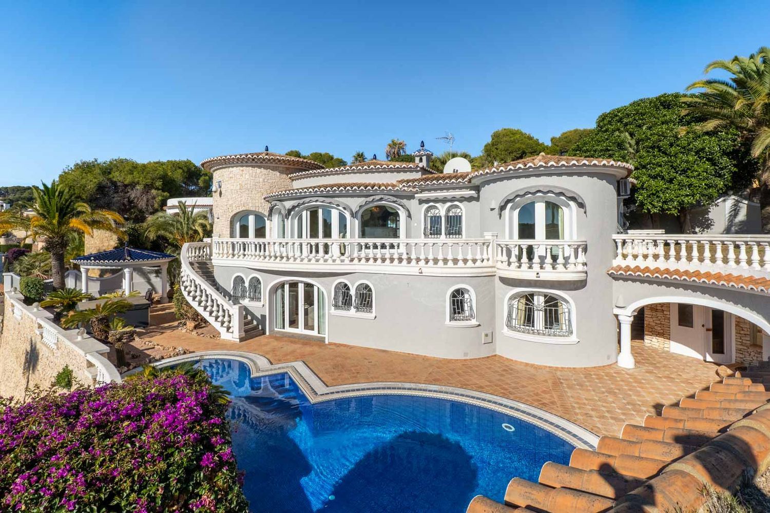 The villa's multi-level exterior features a grey facade, white balustrades, and a distinctive private pool. A curved staircase connects the upper balcony to the terracotta-tiled pool deck, surrounded by Mediterranean flora.