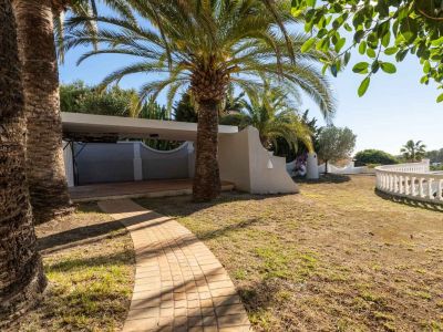 A paved pathway winds through a garden with mature palm trees and green lawn. A covered carport and a white curved balustrade are visible, set against Mediterranean flora under a clear blue sky.