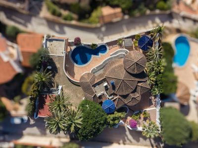 Aerial view of the villa showcasing its tiered terracotta tiled roofs, an irregular-shaped private pool, and a terracotta patio. Surrounded by palm trees and Mediterranean foliage.