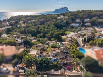 Aerial view showcasing the villa's distinctive tiled roofs amidst lush Mediterranean gardens. The panoramic sea view with the iconic Peñón de Ifach dominates the background.