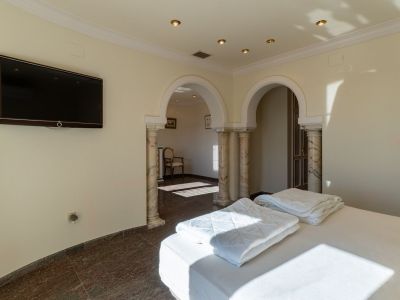 Bedroom featuring a bed, dark marble flooring, and a wall-mounted TV. Two arched passages with decorative marble columns open to an adjacent room, illuminated by recessed lights and natural sunlight.