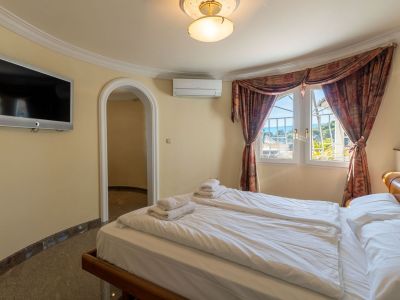 Bright bedroom featuring twin beds, a wall-mounted TV, and partial sea views through windows with patterned curtains. Includes air conditioning and an arched doorway.
