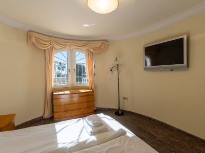 Bright bedroom with a bed, light wood chest of drawers under a curved window, and wall-mounted TV. Dark tiled floor and light walls are bathed in natural light, with a glimpse of outdoor greenery.
