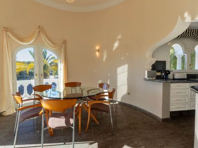 Bright dining area featuring a glass table and wooden chairs, connected to a kitchen bar with arched windows, and offering views of palm trees through French doors.