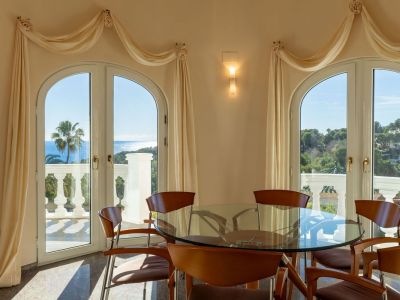Bright dining area featuring a glass table and wooden chairs, framed by arched doors opening to panoramic Mediterranean sea and coastal views.