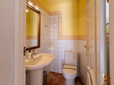 Compact bathroom featuring a pedestal sink, a wooden-framed mirror with overhead lighting, and white tiled walls with decorative floral borders. It includes a toilet and terracotta floor tiles.