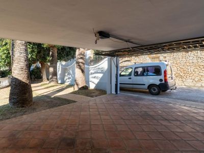 Covered parking area with terracotta tiled flooring, bordered by mature palm trees and a white van parked. A stone wall is visible in the background.