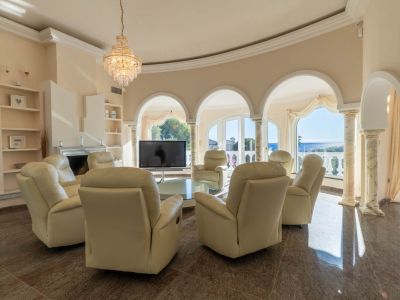 Curved living room featuring recliners, polished marble flooring, and sea views through arched windows. A central crystal chandelier and classic columns enhance the elegant space.