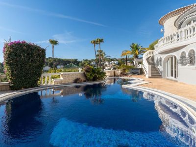 Expansive private pool with blue mosaic tiles and an irregular shape, surrounded by light paving and Mediterranean flora. A grand curved staircase with white balustrades leads to the villa.