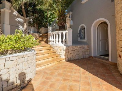 Exterior entrance featuring terracotta tiled flooring and ascending steps. A classical white balustrade and natural stone walls frame the grey facade with an arched doorway.