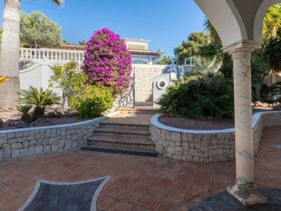 Exterior patio featuring terracotta tiles with a dark mosaic inlay, leading to decorative steps, a stone wall, and a gate adorned with vibrant purple bougainvillea. A marble column frames the view.