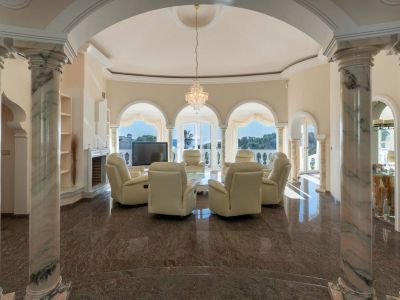 Grand living room featuring polished dark marble floors and elegant marble columns. Arched windows offer bright natural light and views, complementing the central seating area with recliners and a crystal chandelier.