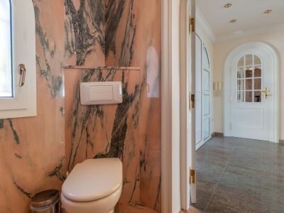 Guest toilet featuring pink veined marble walls and a window, connecting to a hallway with dark tiled flooring and an arched door.