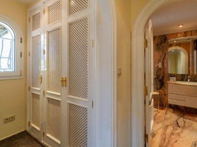 Hallway featuring white louvered built-in wardrobes with gold handles and an arched window. An arched doorway leads to a bathroom with veined marble finishes.