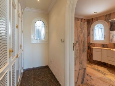 Hallway featuring white louvered wardrobes with gold handles, leading to a bathroom clad in pink marble with two arched windows.
