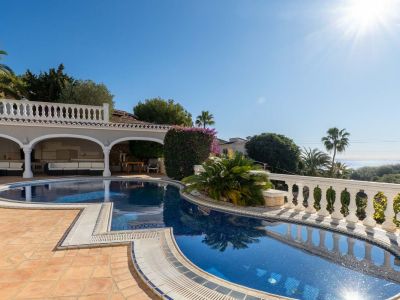 Irregularly shaped blue mosaic pool with terracotta tiling, adjacent to a covered arched terrace with white outdoor sofas. Distant sea views visible through lush greenery.