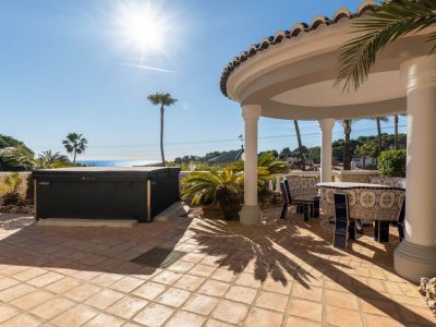 Outdoor terrace featuring a hot tub and a covered dining area with white columns and a tiled roof. A blue and white tiled table and chairs sit on terracotta flooring, offering sea views.