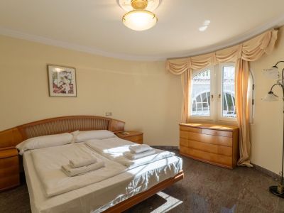 Spacious bedroom featuring a double bed with a wooden headboard, matching nightstands, and a dresser. Natural light enters through a curved window, illuminating the dark tiled floor.