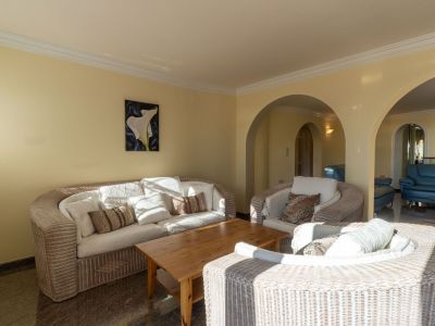 Spacious living area with wicker sofas, light cushions, and a wooden coffee table on dark marble flooring. Elegant arches connect to an adjacent room, bathed in natural light.