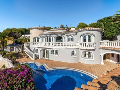 The villa's multi-level exterior features a grey facade, white balustrades, and a distinctive private pool. A curved staircase connects the upper balcony to the terracotta-tiled pool deck, surrounded by Mediterranean flora.