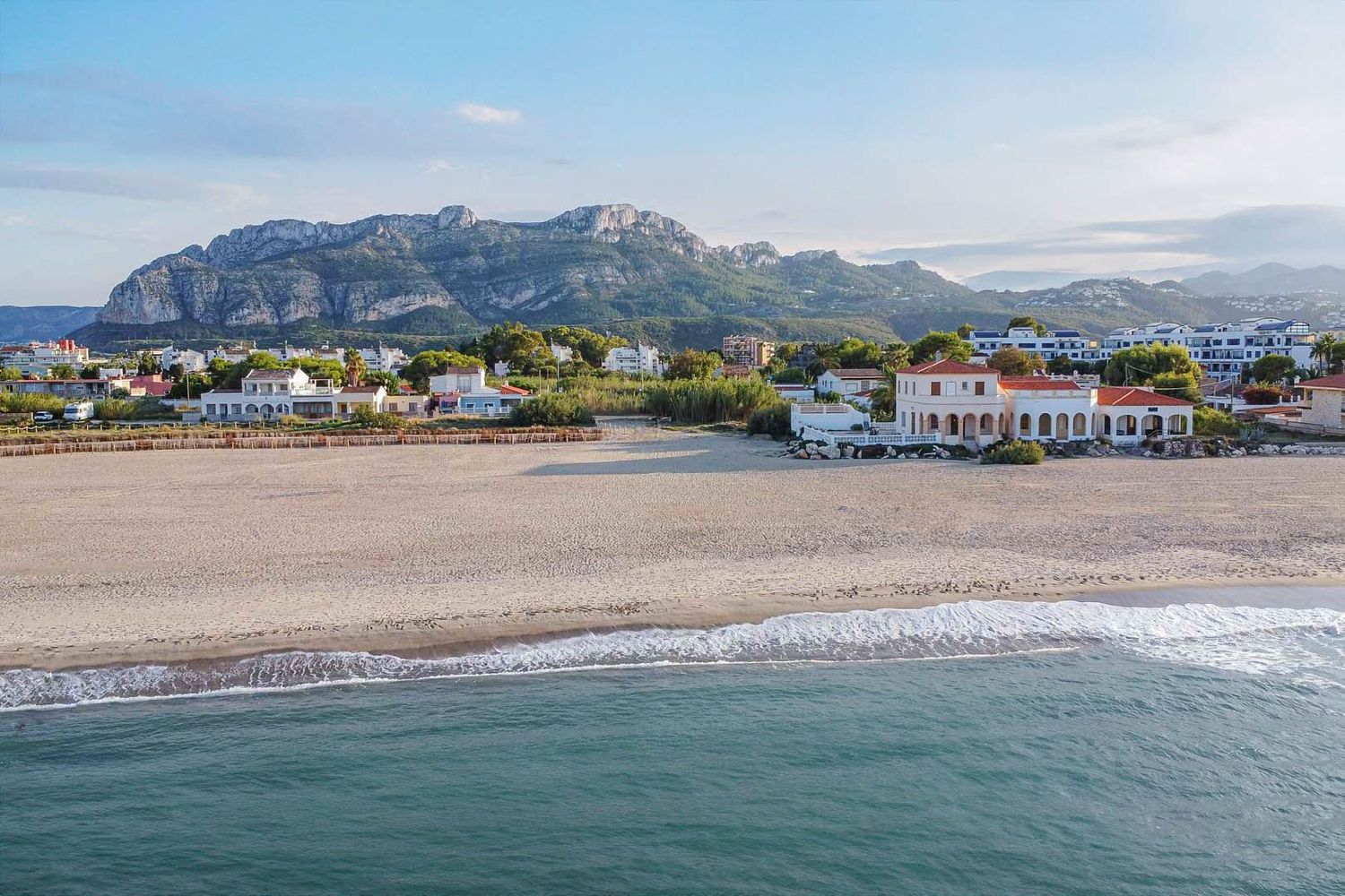 Aerial view of the sandy beach with gentle waves, featuring a classic coastal house with arches, set against the backdrop of the Montgó mountain range.