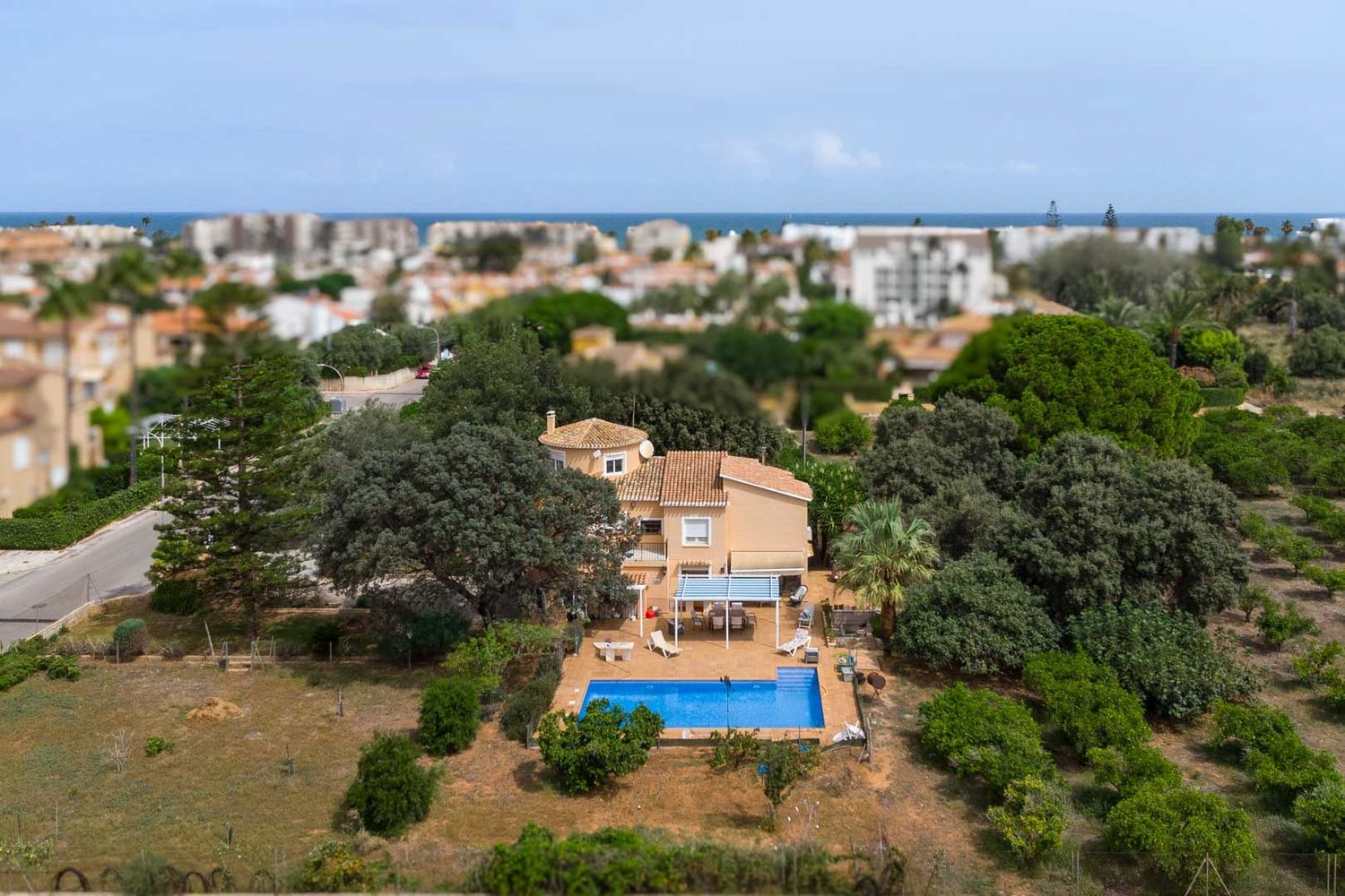 Aerial view of the villa featuring a blue swimming pool, a shaded terrace with an awning, and extensive mature gardens. The Mediterranean Sea is visible in the distance.