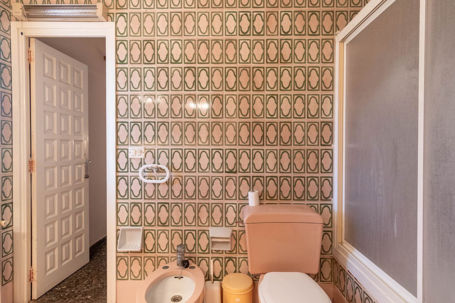 Bathroom with green and beige patterned tiles, pink toilet and bidet, frosted shower screen, and a speckled terrazzo floor.