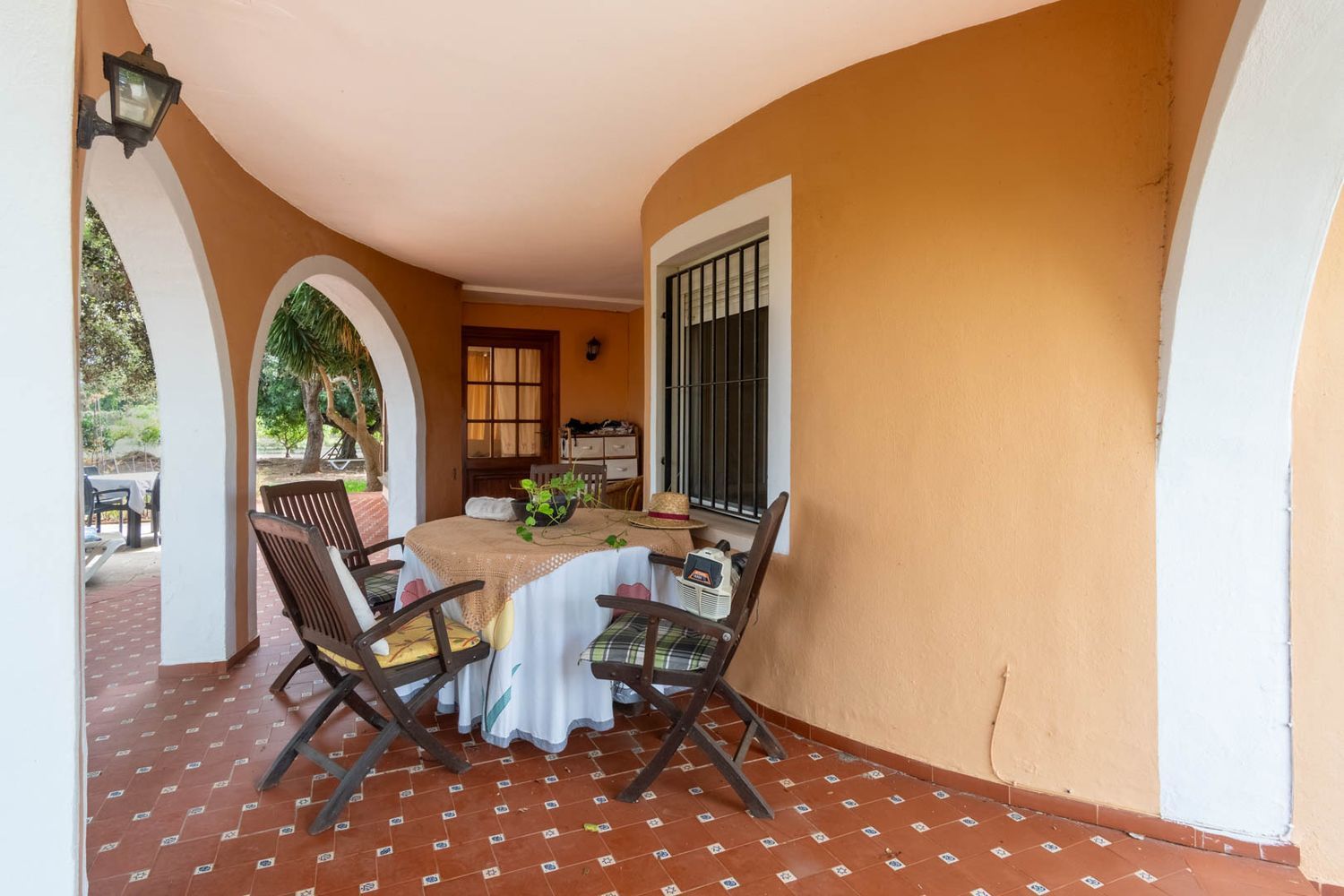 Covered terrace featuring reddish-brown tiled flooring, white arched openings to the garden, and an orange curved wall. Furnished with a round table and wooden chairs.