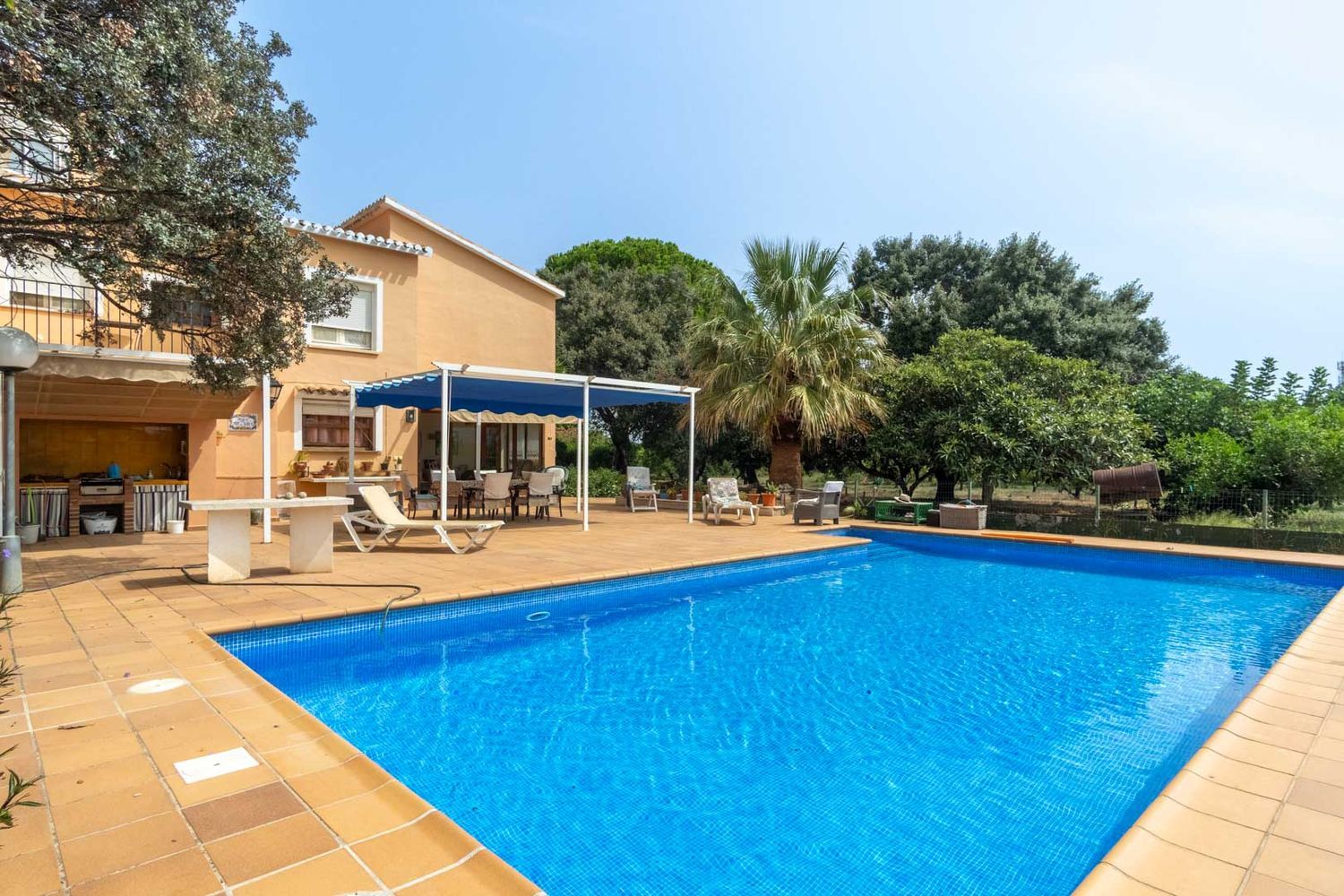 Spacious blue mosaic pool surrounded by a tiled sun terrace, featuring a shaded outdoor dining area and a built-in barbecue zone.