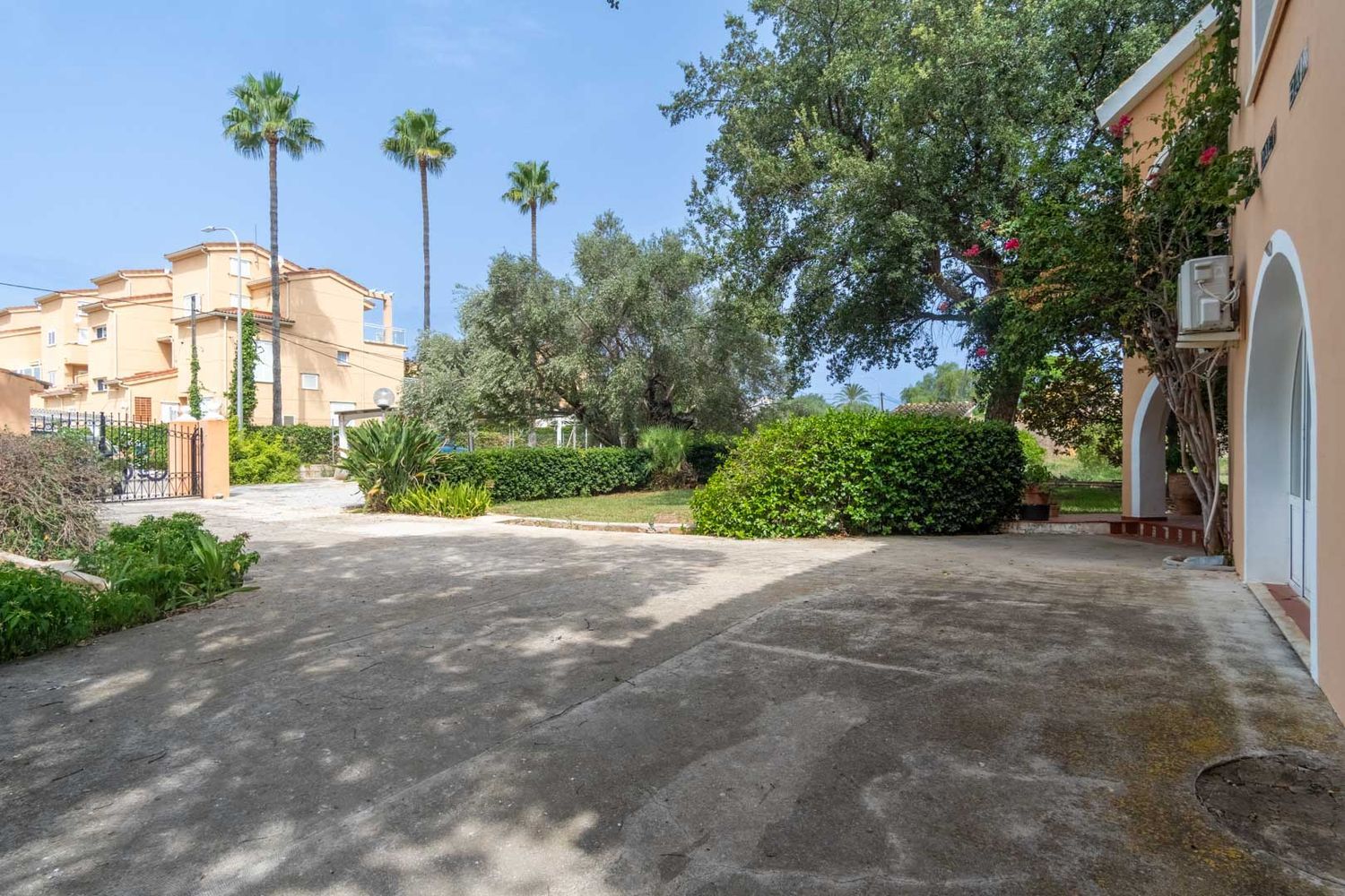 Spacious concrete private parking area, bordered by a Mediterranean garden featuring palm trees, olive trees, and lush shrubs. Part of the chalet's arched facade is visible.
