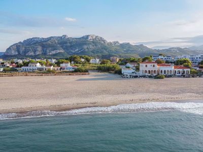 Aerial view of the sandy beach with gentle waves, featuring a classic coastal house with arches, set against the backdrop of the Montgó mountain range.
