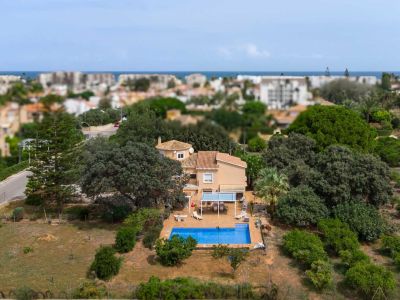 Aerial view of the villa featuring a blue swimming pool, a shaded terrace with an awning, and extensive mature gardens. The Mediterranean Sea is visible in the distance.