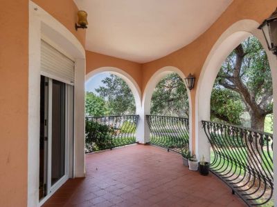 Arched covered terrace featuring terracotta floor tiles and decorative wrought iron railings, providing views of the surrounding garden with mature trees.