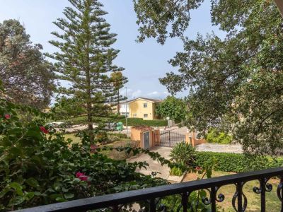 Balcony view framed by a wrought iron railing and vibrant bougainvillea, overlooking a Mediterranean garden with an access path and a yellow house in the background under a clear sky.