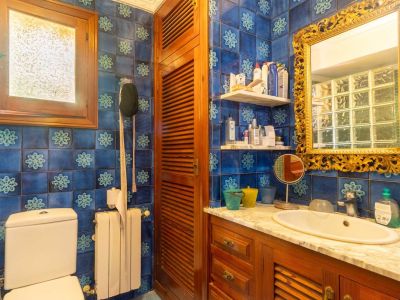 Bathroom featuring distinctive blue patterned tiles, a wooden vanity with white marble top, and an ornate gold-framed mirror. A frosted wooden window provides natural light.