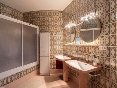 Bathroom featuring distinctive green and beige patterned wall tiles, a double vanity with two pedestal sinks, dual round mirrors with integrated lighting, a shower cabin, and a tall storage cabinet.